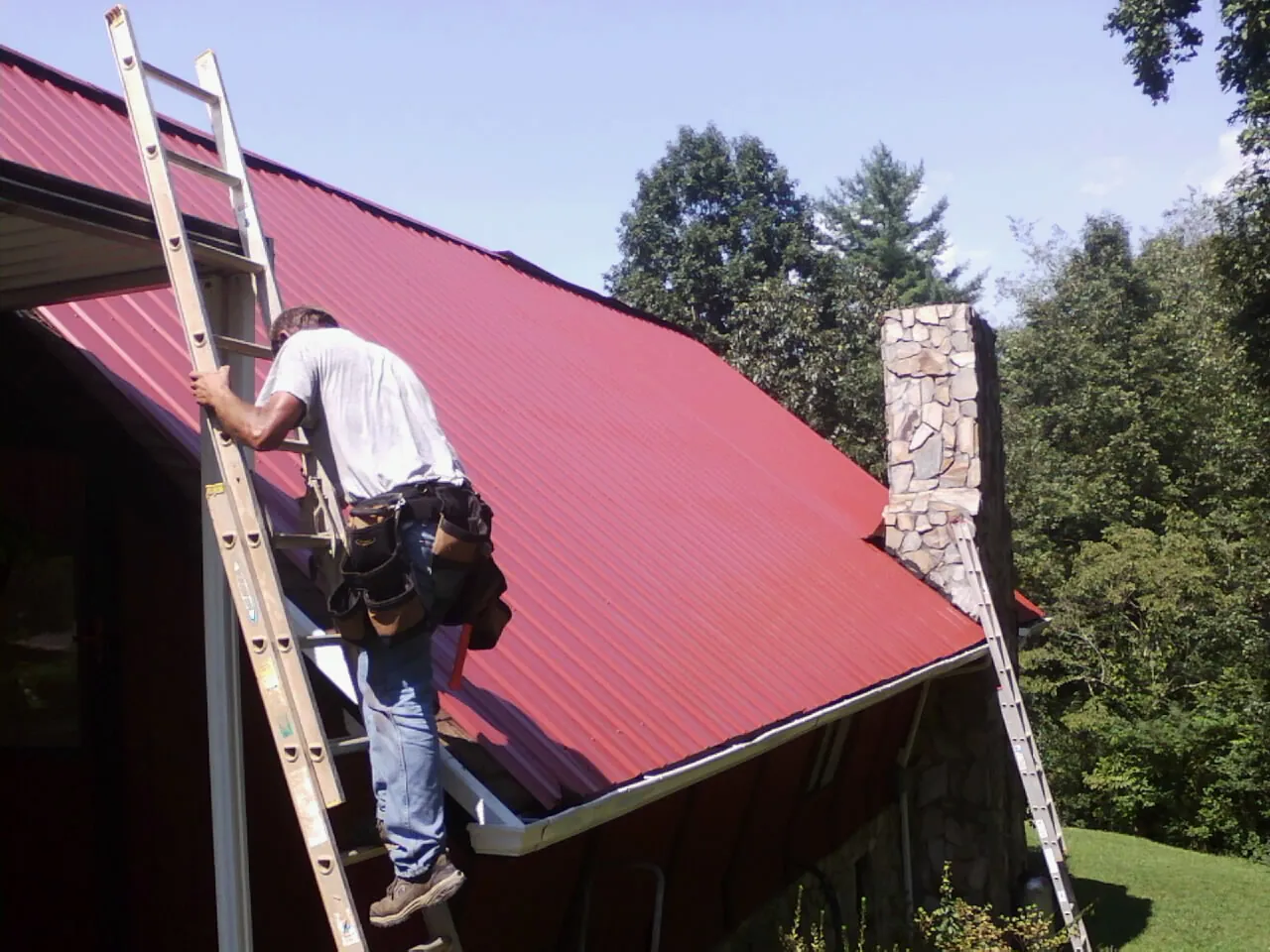 Roofer on a ladder beside a red metal roof and stone chimney — Soffit & Fascia Repair by Roofing Brothers in Hendersonville